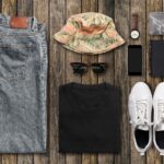 Image of folded clothing, including a pair of blue jeans, a summer hat, sunglasses, a black t-shirt, white trainers, and accessories.