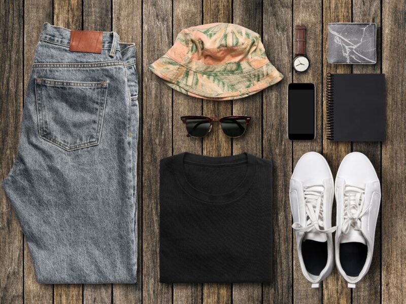 Image of folded clothing, including a pair of blue jeans, a summer hat, sunglasses, a black t-shirt, white trainers, and accessories.