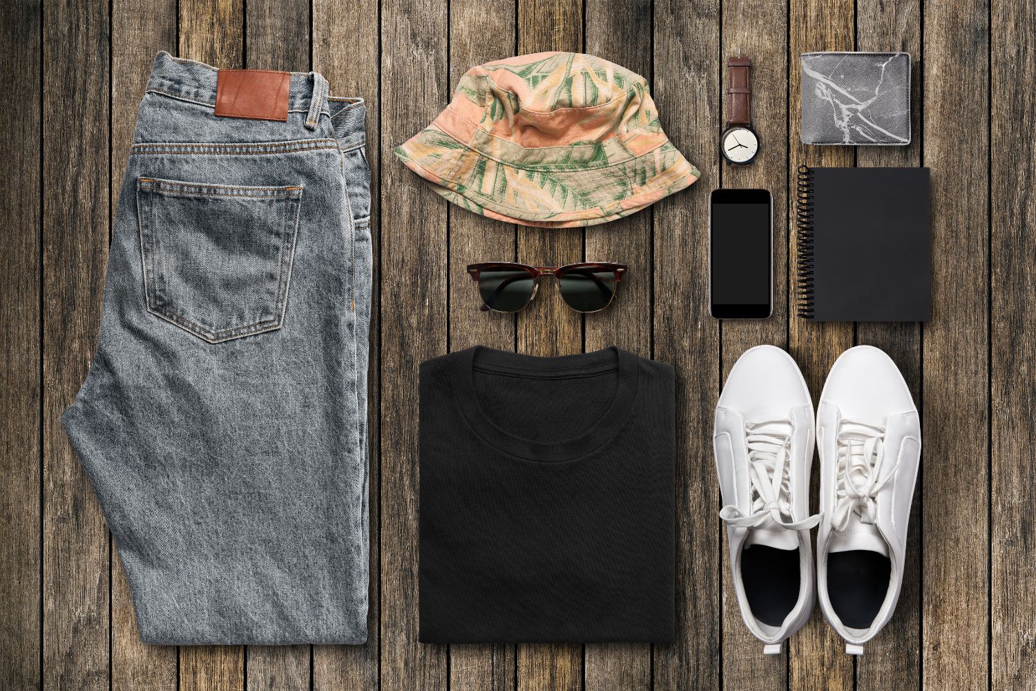 Image of folded clothing, including a pair of blue jeans, a summer hat, sunglasses, a black t-shirt, white trainers, and accessories.