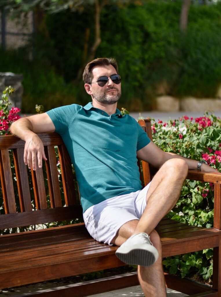 Man sitting down on a bench wearing a turquoise polo shirt and a light beige chino shorts.